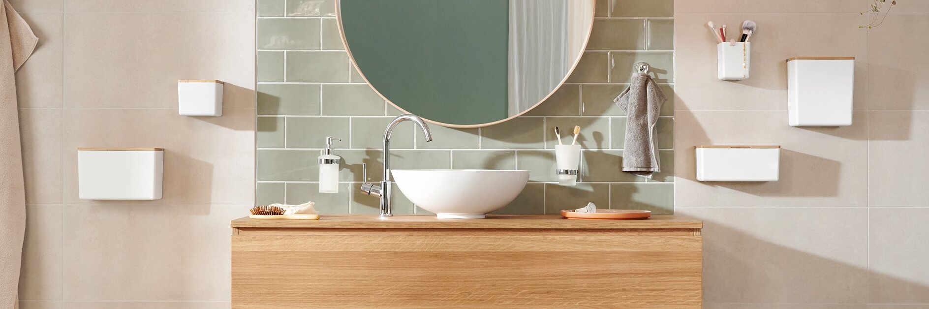 Modern bathroom with a wooden vanity and round mirror above. A white sink and silver faucet are centered on the vanity. Green tiled wall, pendant light, and assorted toiletries are visible. Neutral-toned walls and floor create a calming atmosphere, showcasing the seamless application of tesa tape for all mounting needs. (This text has been generated by AI)