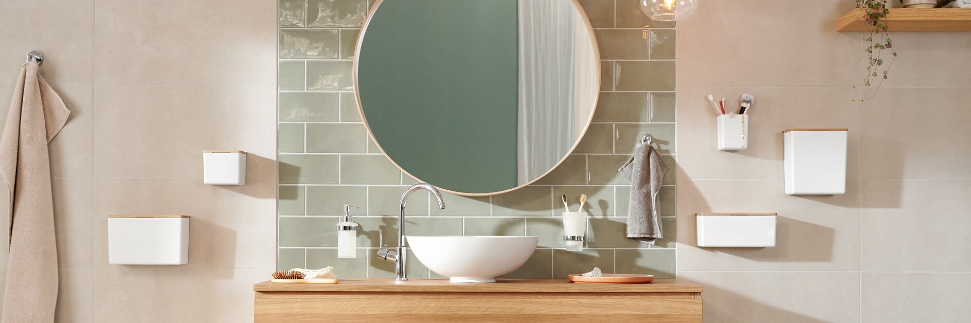 A bathroom features a round mirror above a wooden vanity with a white sink. Green tiles serve as a backsplash. Several white shelves and a towel holder are mounted on the wall using tesa tape. A hanging light fixture and a beige towel are visible. (This text has been generated by AI)