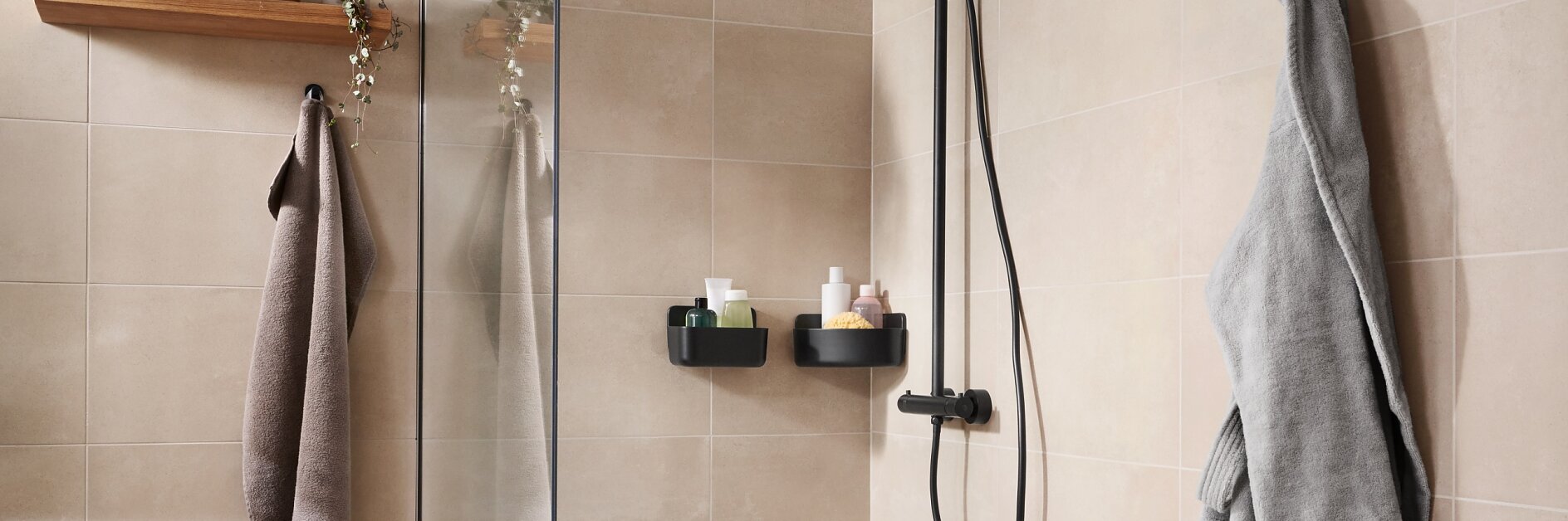 A bathroom with beige tiles features a glass shower enclosure and a black rain showerhead. Two wall-mounted shelves hold toiletries secured with tesa tape. A wooden shelf with hanging greenery is outside the shower, attached using tesa tape. Two towels are hung nearby with the help of tesa tape. (This text has been generated by AI)