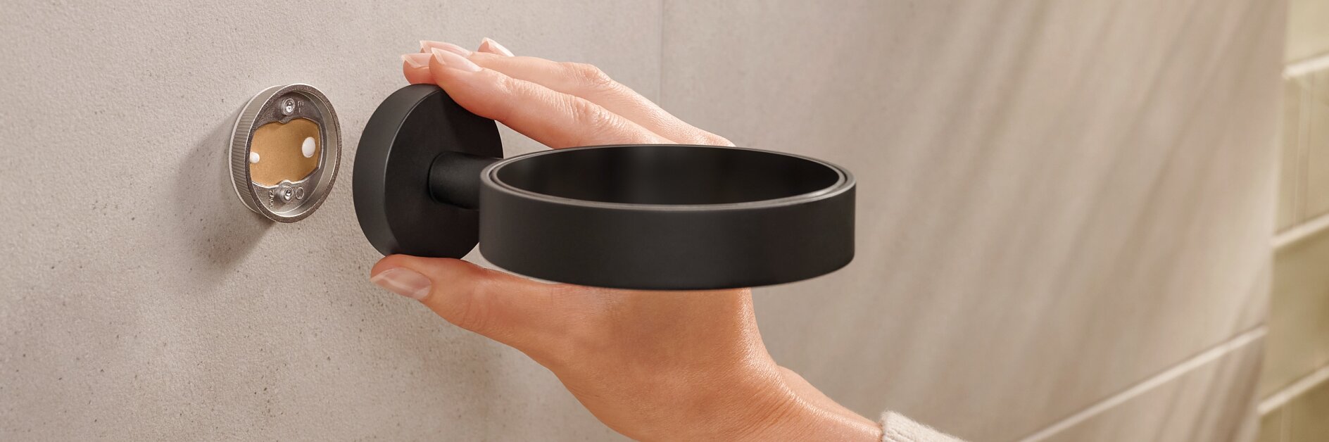 A person is installing a black wall-mounted holder onto a beige tiled wall. The holder is circular, and the persons hand is seen adjusting it, aligning it with the mounting fixture using tesa tape. (This text has been generated by AI)