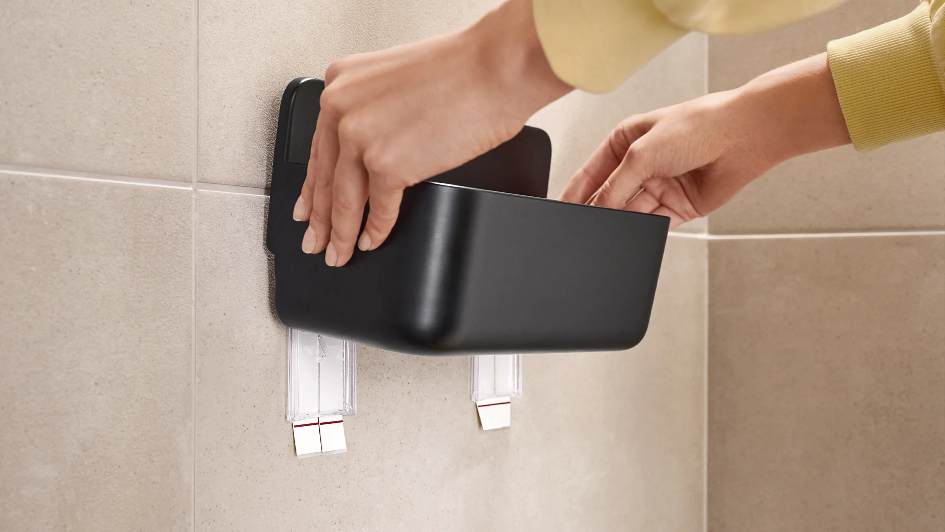 Hands are attaching a black container to a beige tiled wall using two strips of tesa tape. The container appears to be a wall-mounted organizer or storage shelf. (This text has been generated by AI)