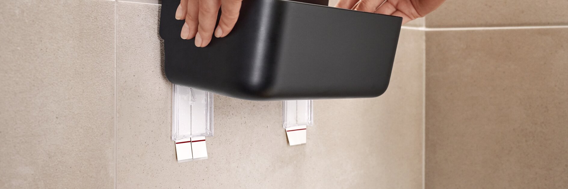 Hands are attaching a black container to a beige tiled wall using two strips of tesa tape. The container appears to be a wall-mounted organizer or storage shelf. (This text has been generated by AI)