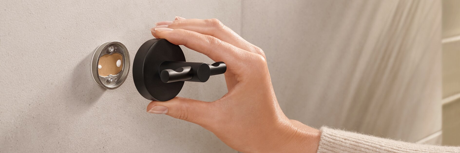 A hand wearing a beige sweater is attaching a round black fixture onto a circular base on a light gray tiled wall using tesa tape. A metal mounting bracket is partially visible next to it. (This text has been generated by AI)