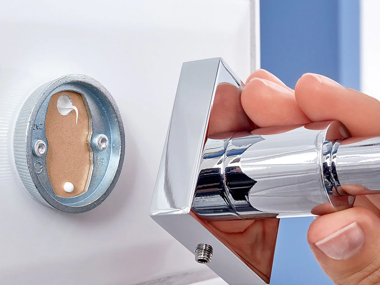 A hand is holding a shiny chrome door handle against a white door. The handle is partially detached, revealing the mounting plate and screws inside the door. The image focuses on the installation or repair process of the door handle using tesa tape. (This text has been generated by AI)