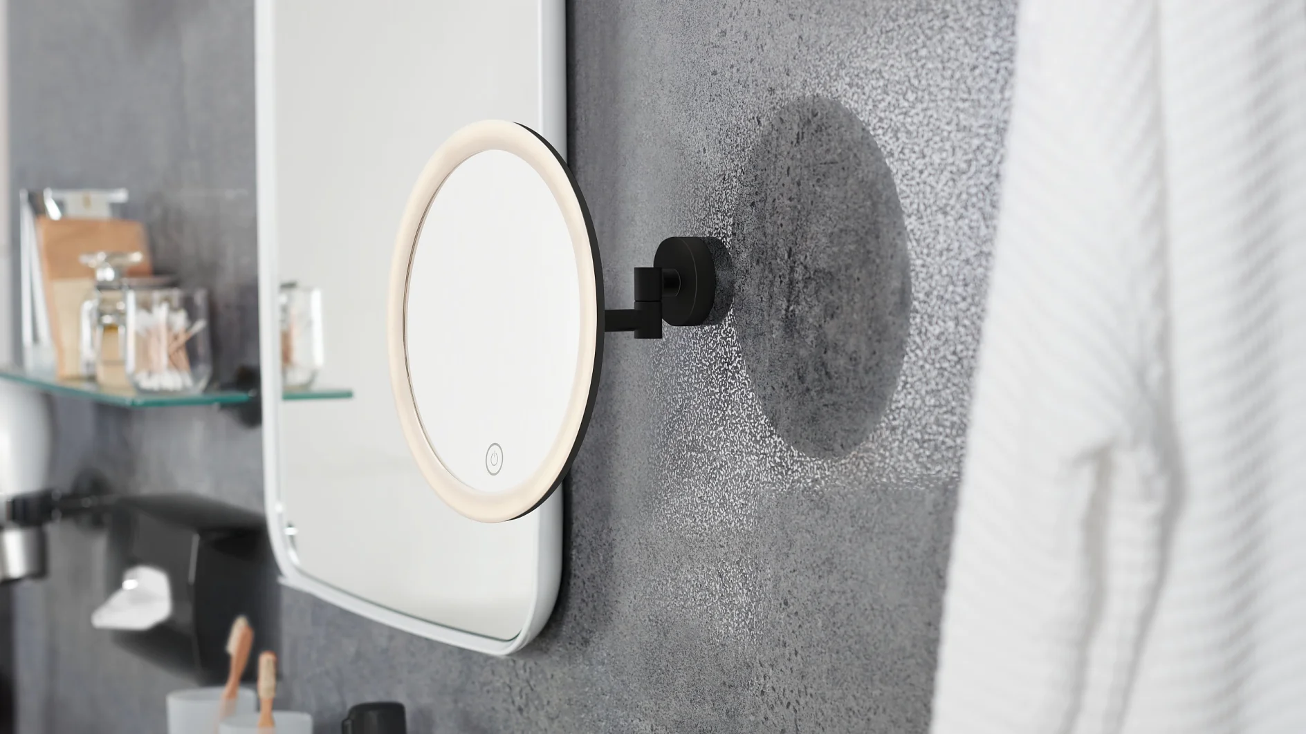 A bathroom wall features a circular, lighted mirror attached to an adjustable arm. Nearby, shelves hold jars and toiletries secured with tesa tape. A white bathrobe hangs on the right. The wall has a textured, speckled finish. (This text has been generated by AI)