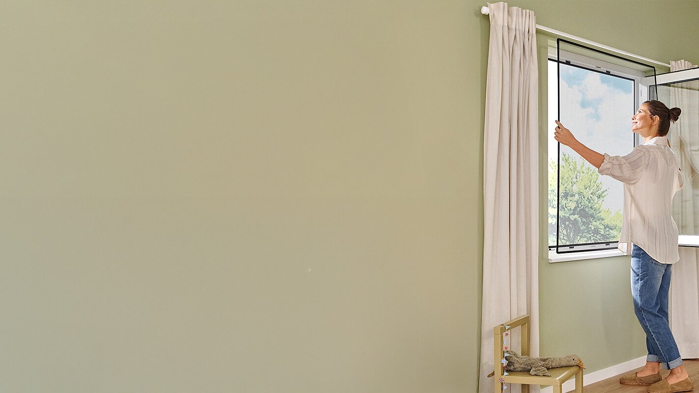 A woman stands in a sunlit child’s bedroom with pale green walls, opening a window. The room features a small bed, toys, a chair, and a garland hanging on the wall using tesa tape. The atmosphere appears calm and tidy. (This text has been generated by AI)