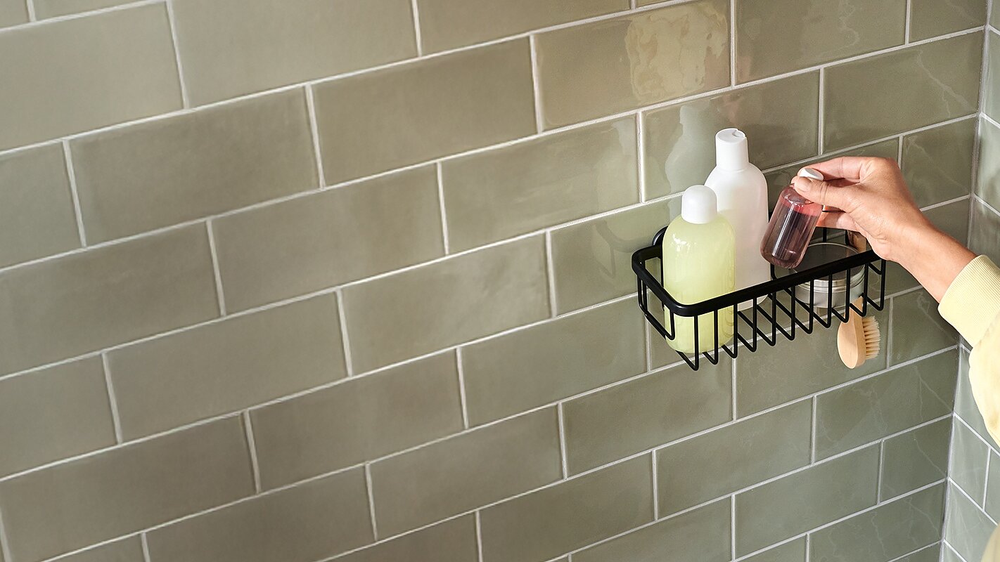A person standing in a bathroom reaches for a small bottle in a black wall-mounted shower caddy, which is secured with tesa tape. The caddy also holds a green bottle, a white pump bottle, and a wooden brush. The wall is covered with light green tiles. (This text has been generated by AI)