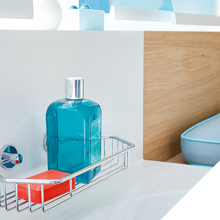 A metal shower caddy is mounted on a white wall using tesa tape. It holds a blue bottle and an orange soap bar. In the background, theres a white shelf, wooden paneling, and blue cushions. An inset image shows a close-up of the empty caddy. (This text has been generated by AI)