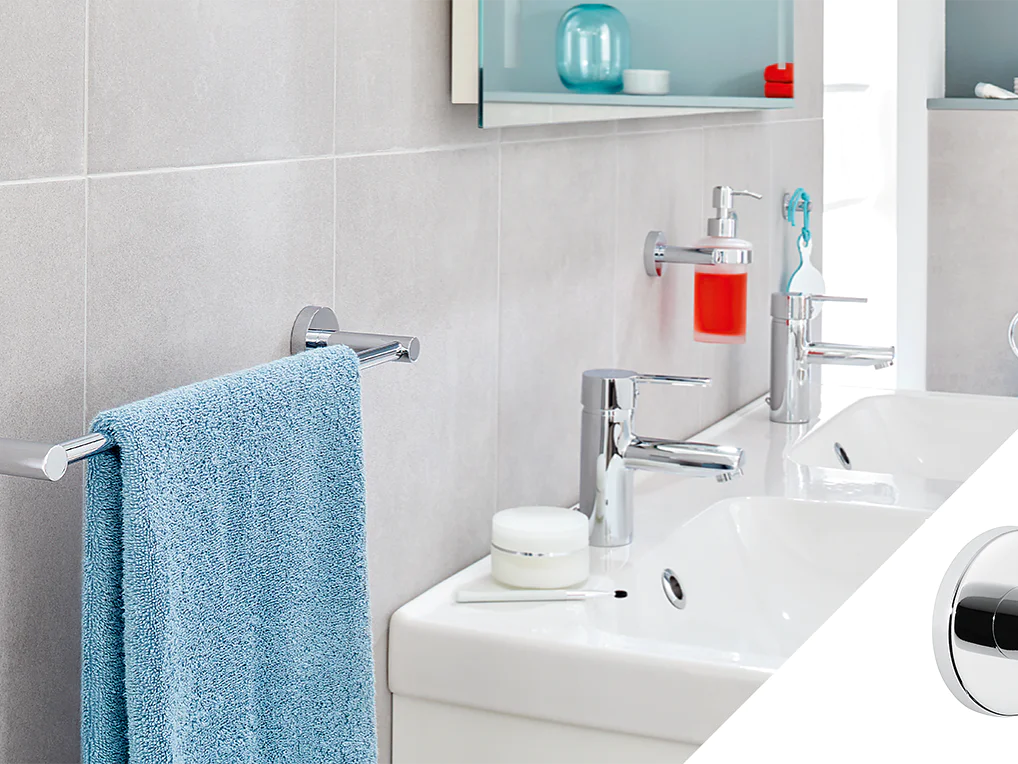 A modern bathroom features two white sinks with chrome faucets, a blue towel on a chrome towel bar, and a red liquid soap dispenser. The inset shows a close-up of a chrome wall hook attached with tesa tape on a light gray tiled wall. (This text has been generated by AI)