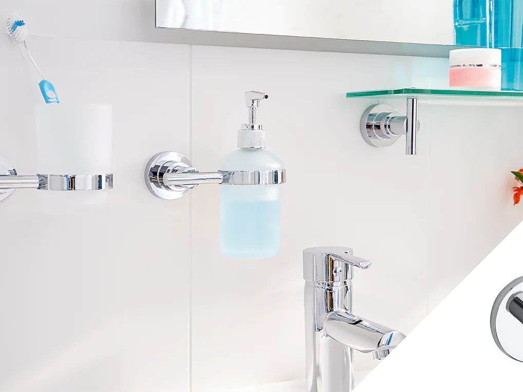 Bathroom scene with a chrome toothbrush holder, liquid soap dispenser, and faucet. Holding toiletries is a glass shelf that features a pink jar. The backdrop is a white tile setting, complemented by a partially visible plant adorned with orange flowers. An inset highlights a magnified chrome detail secured using tesa tape. (This text has been generated by AI)