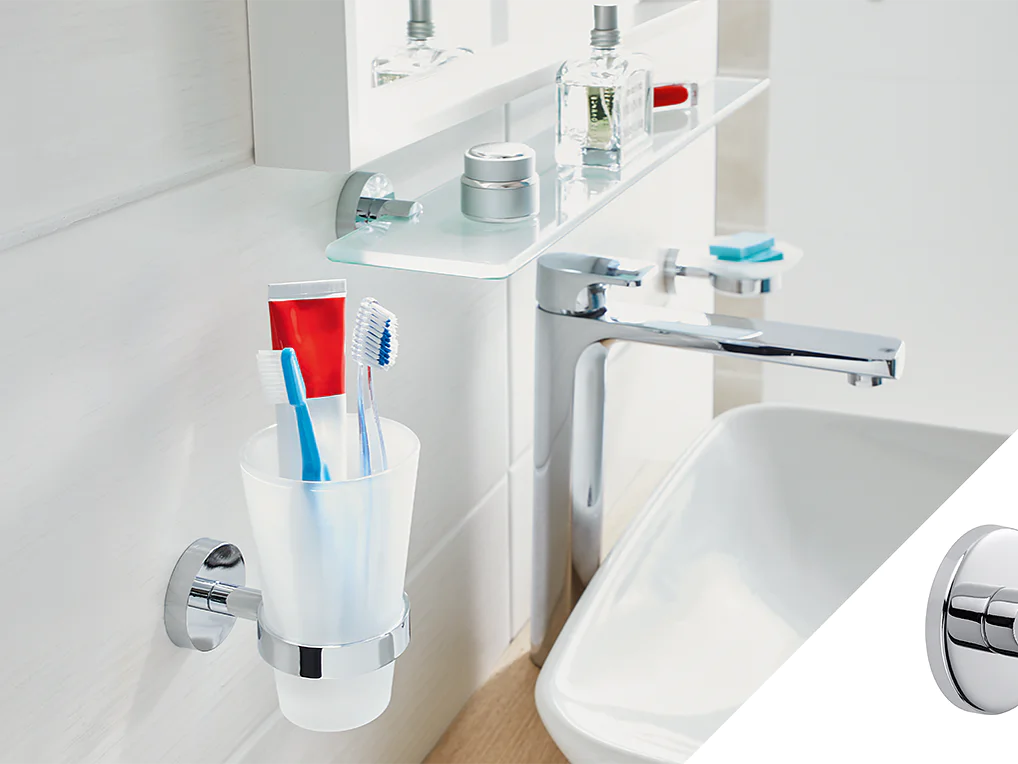 A bathroom sink area features a white cup holding toothbrushes and toothpaste mounted on the wall using tesa tape. Above, a shelf holds toiletries. A silver faucet is attached to the sink. On the right, a silver wall hook is shown. (This text has been generated by AI)