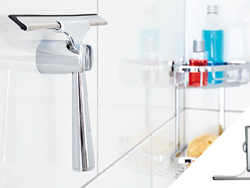 A metallic shower squeegee hangs on a hook attached to a tiled wall using tesa tape. In the background, a shower caddy holds various bottles and toiletries. An inset at the bottom right provides a close-up of the squeegee. (This text has been generated by AI)