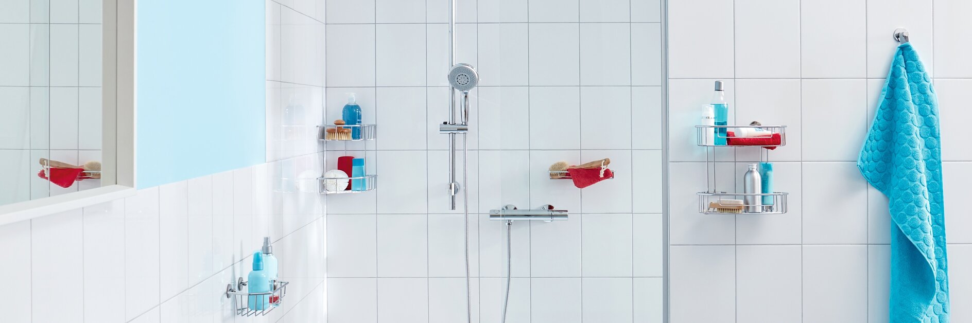 A white tiled bathroom features a shower with a silver handheld showerhead and adjustable bar. There are various toiletries on the corner shelves, and a bright blue towel hangs on the right side with tesa tape. (This text has been generated by AI)