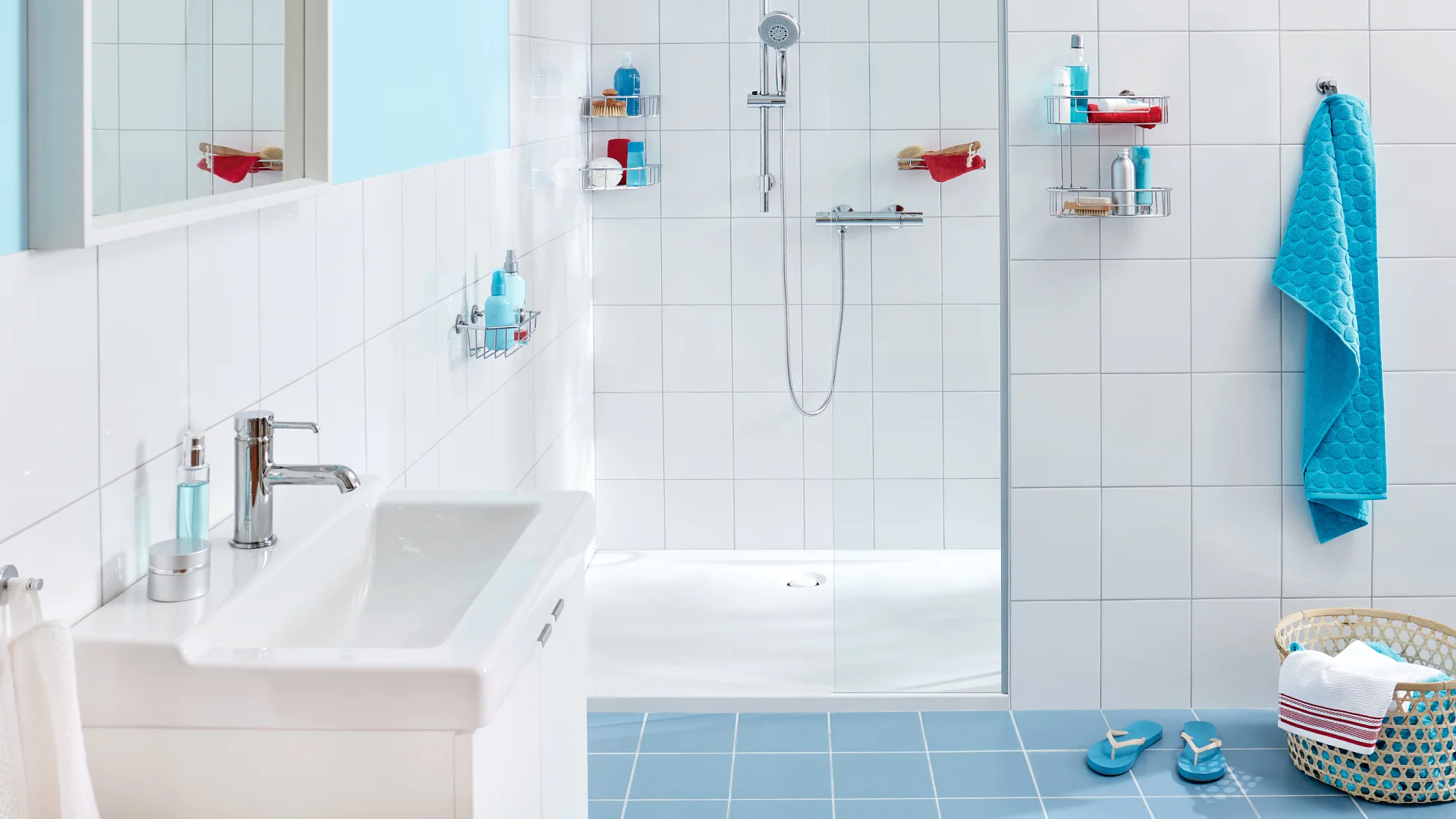 A bathroom with blue and white tiles features a white sink and a walk-in shower. The shower has wall-mounted shelves with toiletries. A basket with a towel and slippers sits on the floor, and a towel hangs next to the sink, secured using tesa tape. (This text has been generated by AI)