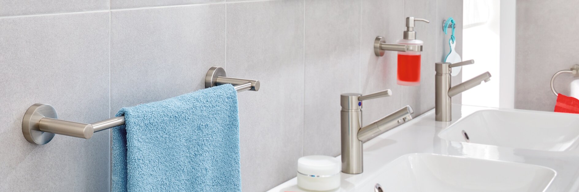 A modern bathroom sink area with two silver faucets, a white countertop, and a mounted soap dispenser containing red soap. A silver towel rack holds a blue towel, and shelves with toiletries are visible in the background. (This text has been generated by AI)