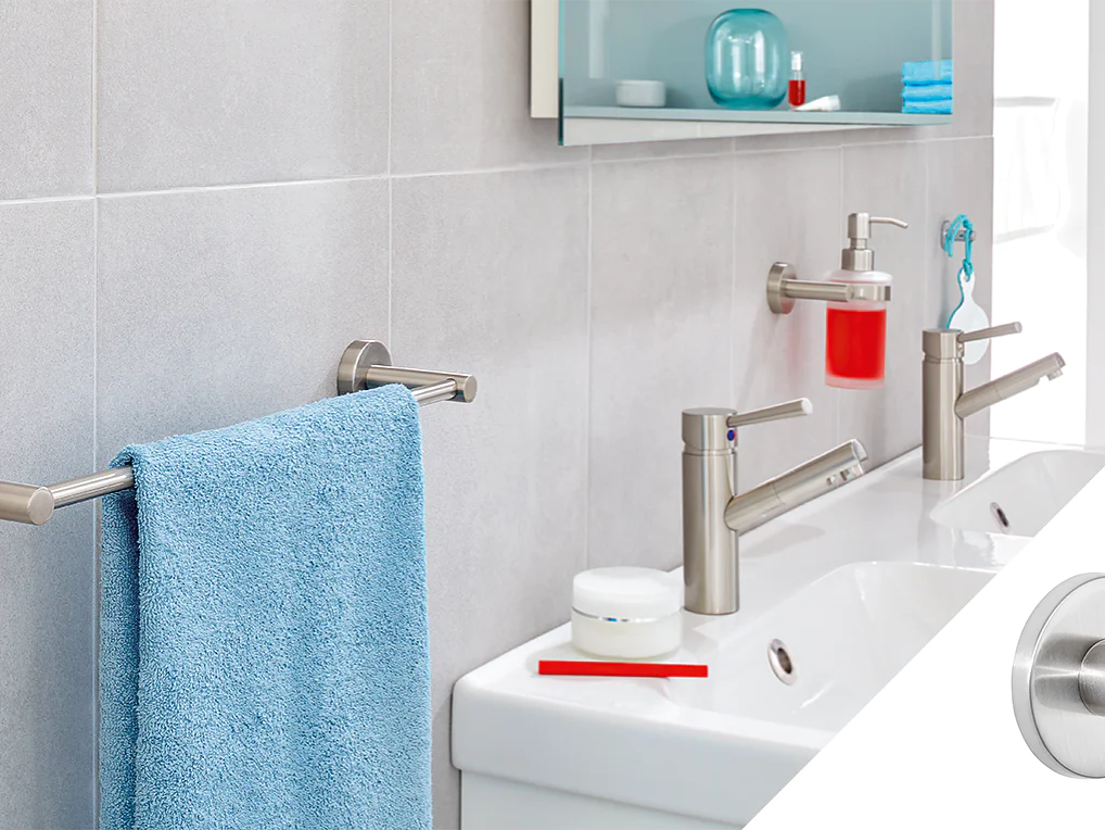 A modern bathroom features a double sink with two silver faucets and a red toothbrush. A blue towel hangs on a silver bar against a light gray tiled wall, secured by tesa tape. A small round silver wall hook is shown separately on the right, also attached using tesa tape. (This text has been generated by AI)