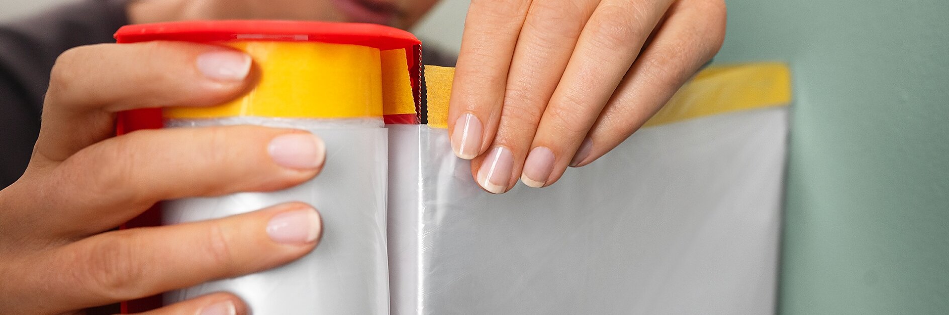 A person unrolls a sheet of clear plastic from a red and yellow roll, preparing to attach it to a surface with tesa tape. Their hands are close to the roll, and the focus is on their actions. (This text has been generated by AI)