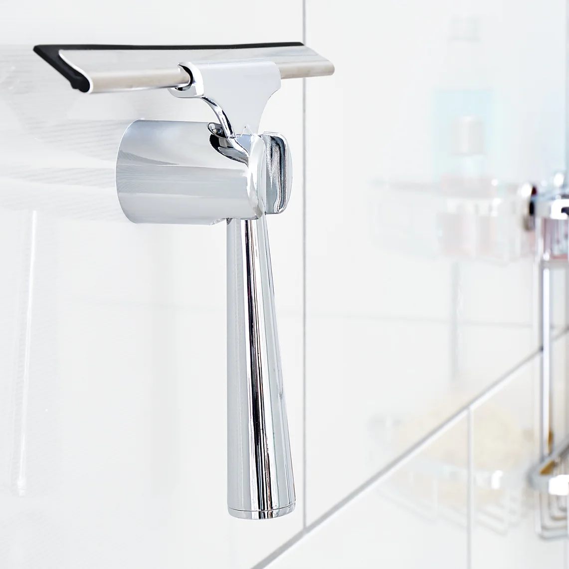 A chrome squeegee hangs on a white tiled bathroom wall, fastened securely with tesa tape. In the background, there are shower caddies holding a blue bottle, a red bottle, and a yellow sponge. (This text has been generated by AI)