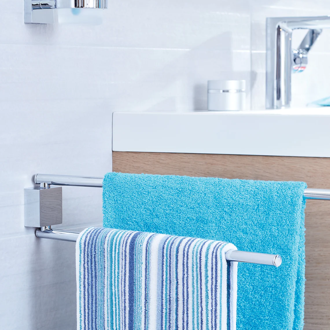 A modern bathroom features a white sink with a chrome faucet, set on a wooden cabinet. In the foreground, a towel rack holds a blue towel and a striped towel, mounted on the tile wall using tesa tape. (This text has been generated by AI)