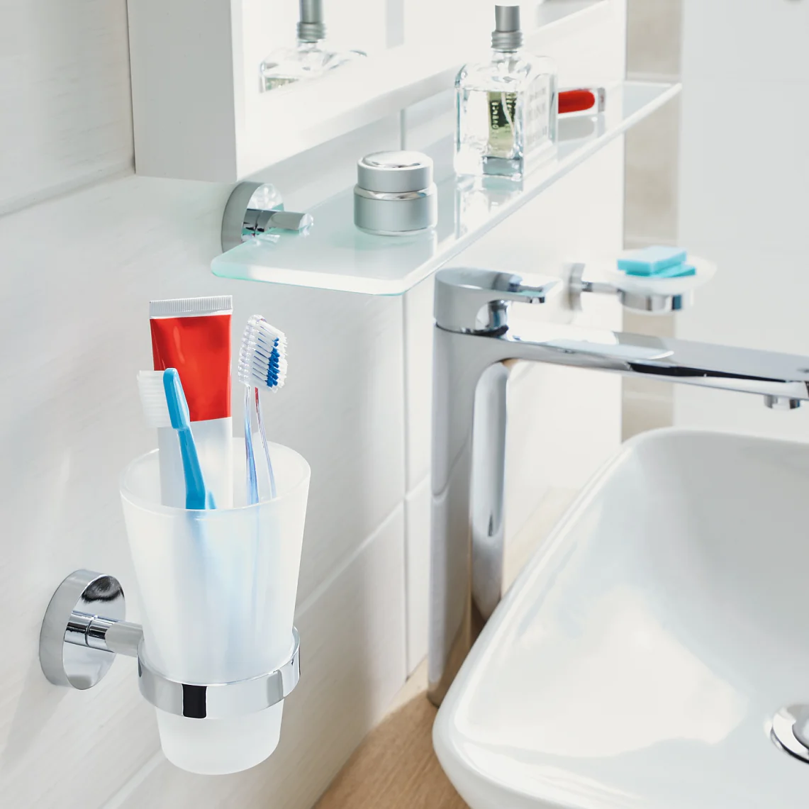 A bathroom sink area featuring a white basin with a silver faucet. A glass shelf above holds toiletries like cologne and creams. A wall-mounted cup contains two toothbrushes and a tube of toothpaste secured neatly in place using tesa tape. The background is white and clean. (This text has been generated by AI)