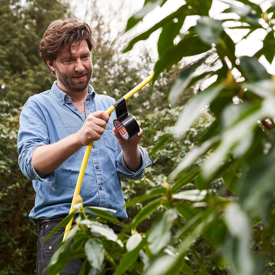 A man wearing a denim shirt is wrapping black tesa tape around a yellow garden hose in a leafy outdoor setting. (This text has been generated by AI)