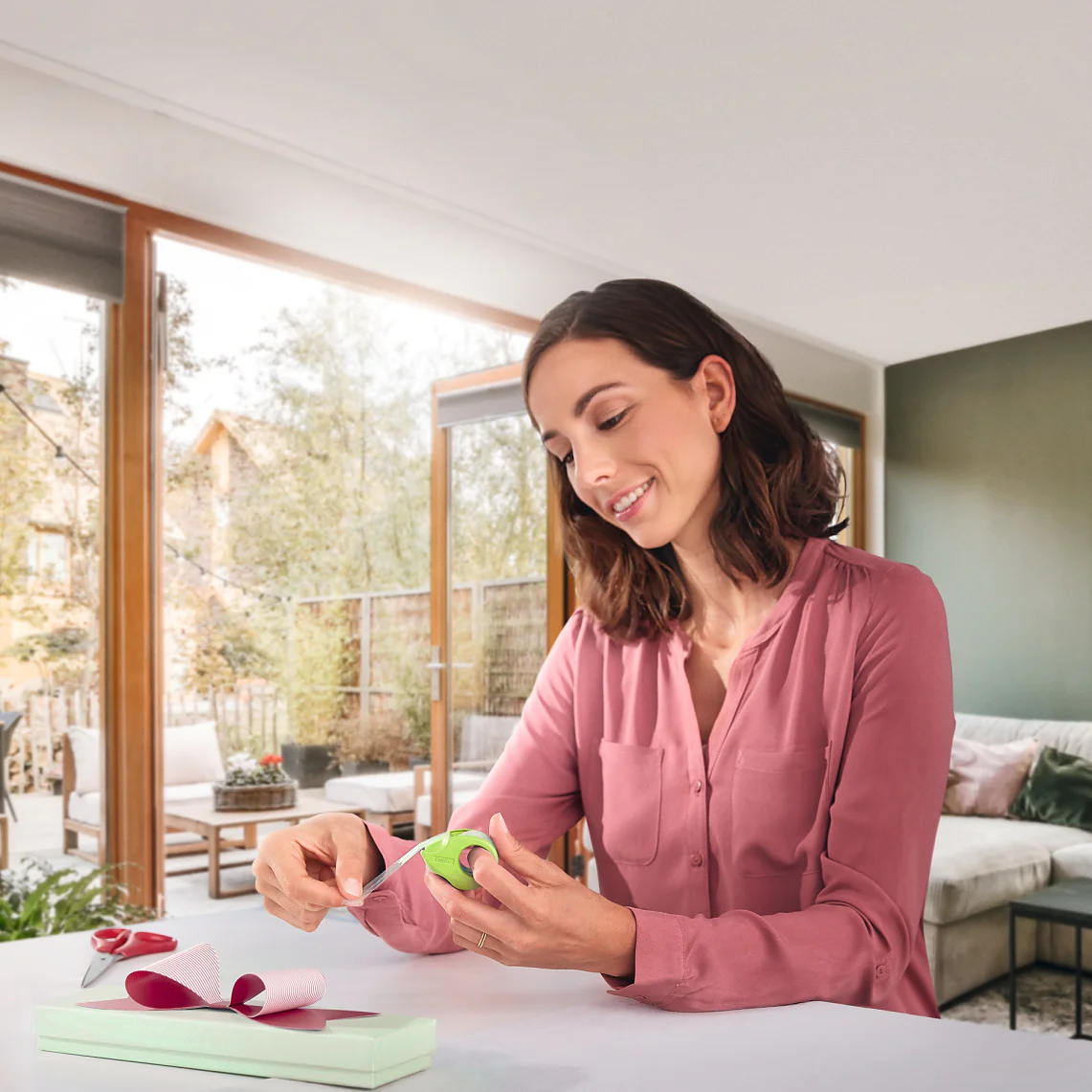 A woman in a pink blouse sits at a table in a modern living room, wrapping a box with green ribbon and tesa tape. Scissors and a spool of ribbon are on the table. Large windows and a patio are visible in the background. (This text has been generated by AI)
