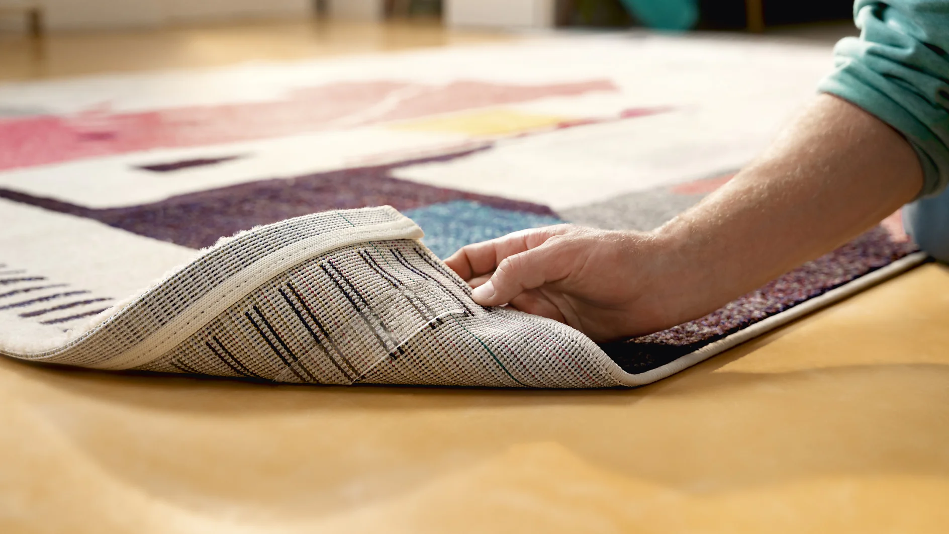 A person lifts the corner of a colorful, geometric-patterned rug, revealing the underside. The rug is placed on a smooth, tan floor using tesa tape. (This text has been generated by AI)