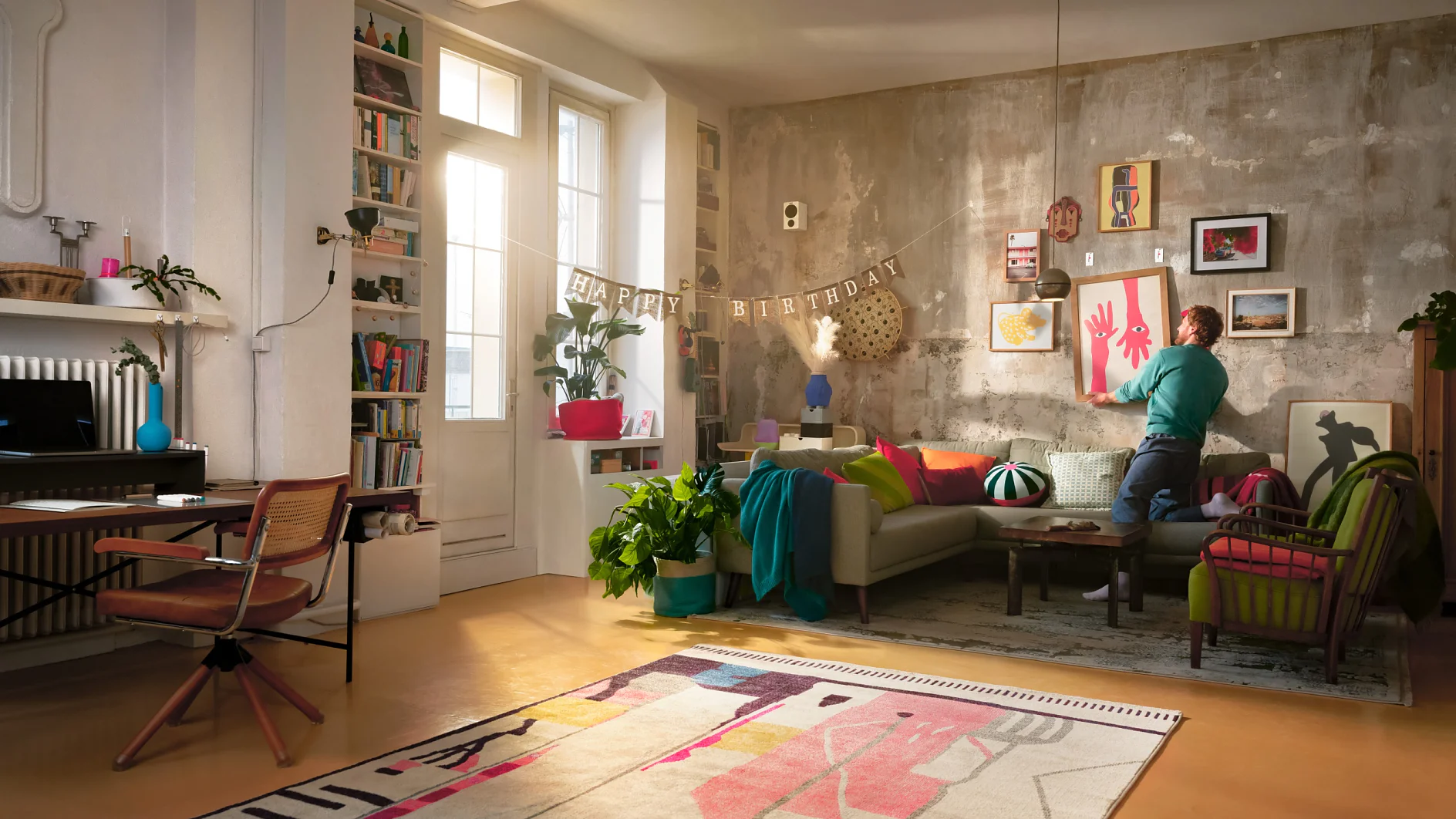 A person stands in a cozy living room, adjusting decorations. The room has colorful art, plants, a couch, and a rug. A Happy Birthday banner hangs above, secured with tesa tape. Light streams through a window beside a bookshelf and desk. (This text has been generated by AI)