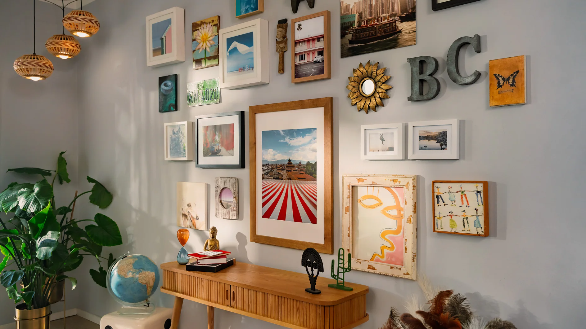 A wall adorned with various framed pictures and art pieces, featuring landscapes and abstract designs, surrounds the letters BC. Below is a wooden console table with decor items such as a globe and a small sculpture. A plant is situated in the corner. The artwork is secured using tesa tape for a clean and professional display. (This text has been generated by AI)