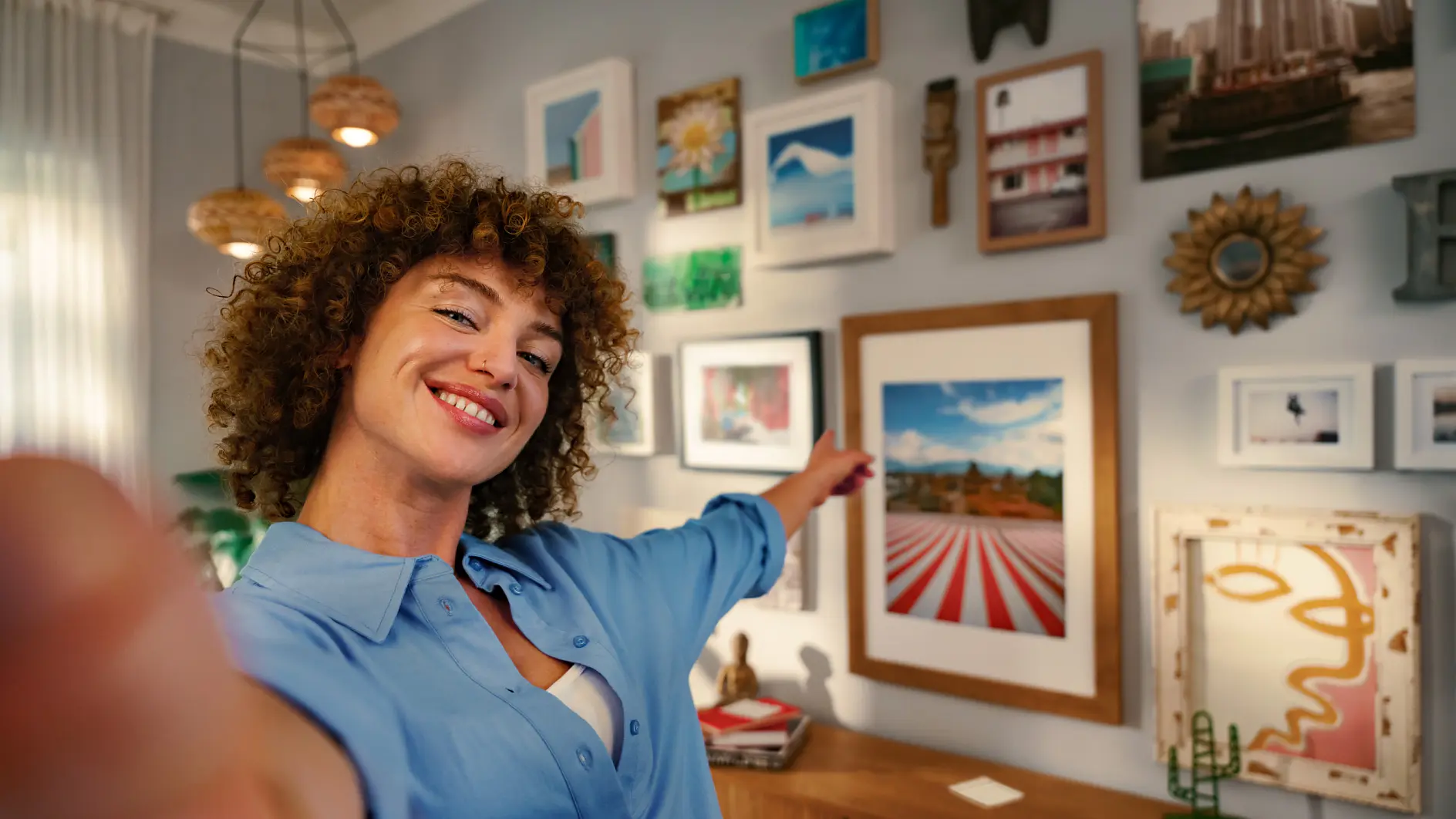 A person with curly hair in a blue shirt takes a selfie while pointing to an art-filled wall. The wall features a variety of framed pictures and art pieces in different sizes and styles. The person is smiling, and the room is warmly lit.