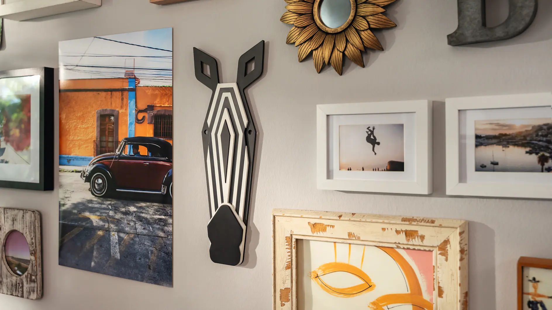A wall displaying a collection of framed photos and art, all securely held with tesa tape. Theres a colorful car photo, a decorative zebra head, a sunflower mirror, and various abstract pieces in different frame styles. The wall is painted light gray.