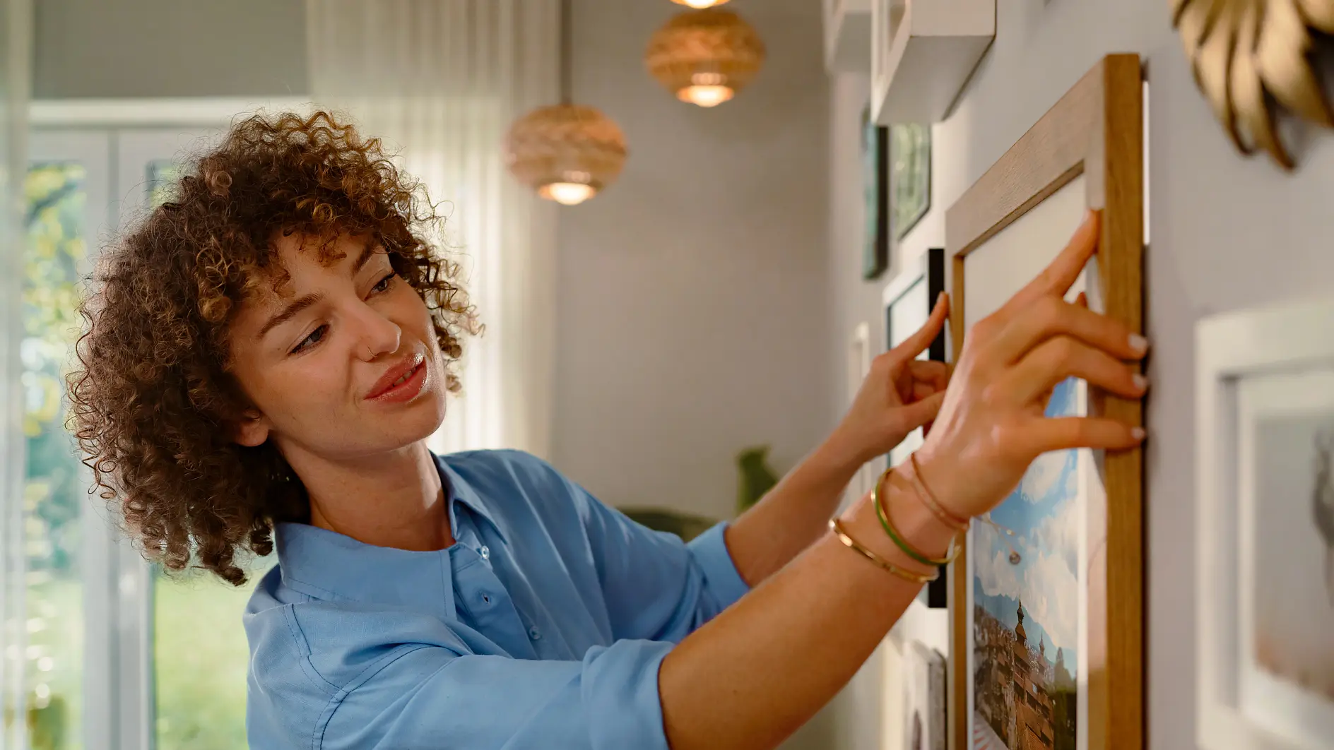 A person with curly hair wearing a blue shirt is hanging a picture on a wall. They are adjusting the frame with both hands using tesa tape. The room has pendant lights and a window with white curtains in the background. (This text has been generated by AI)