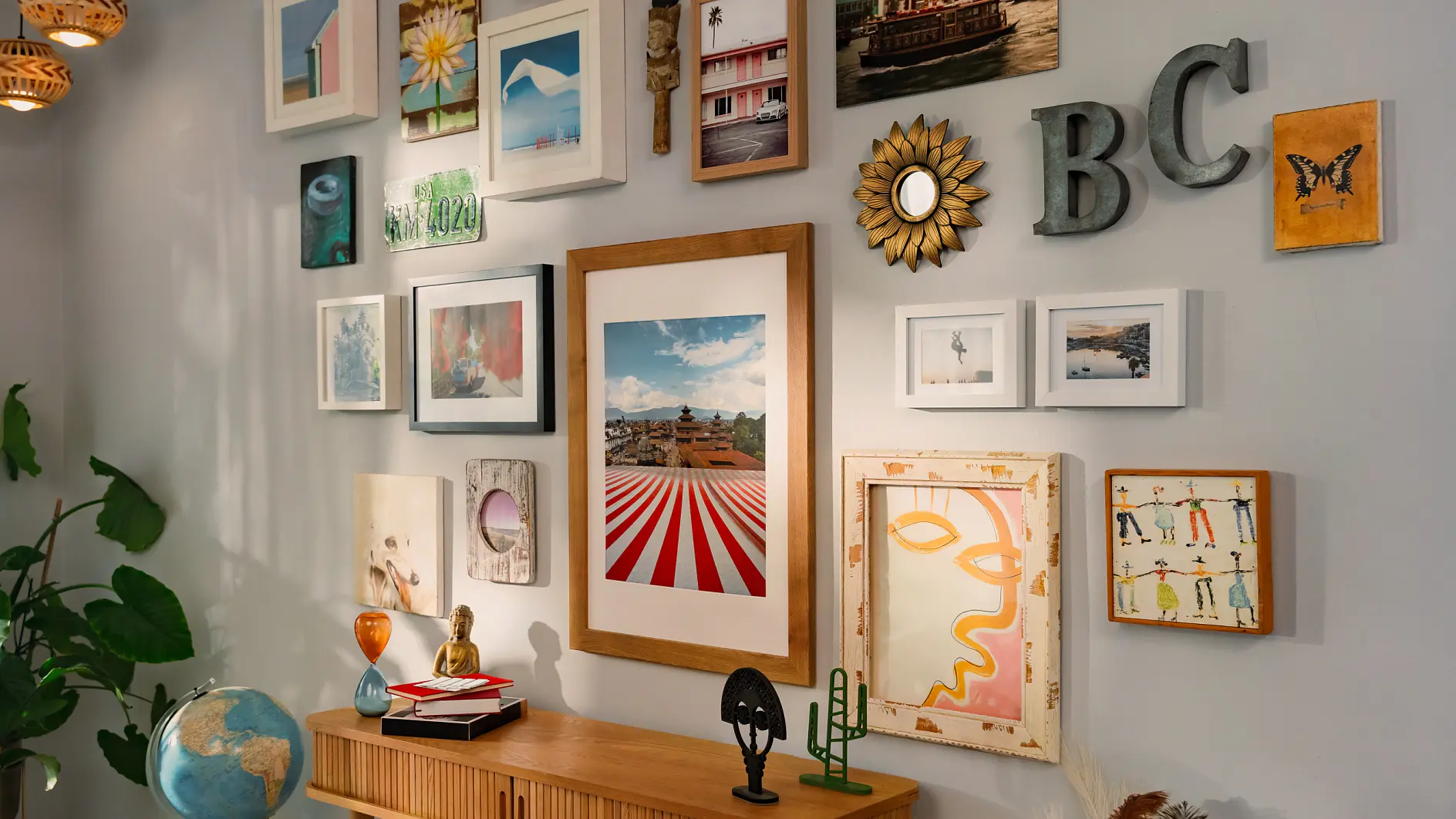 A gallery wall featuring a variety of framed photos and artwork in different styles, alongside decorative letters spelling BC. Below, a wooden console table holds various objects, including a globe and small sculptures. A large plant is visible on the left. All elements are securely mounted using tesa tape for reliable support.