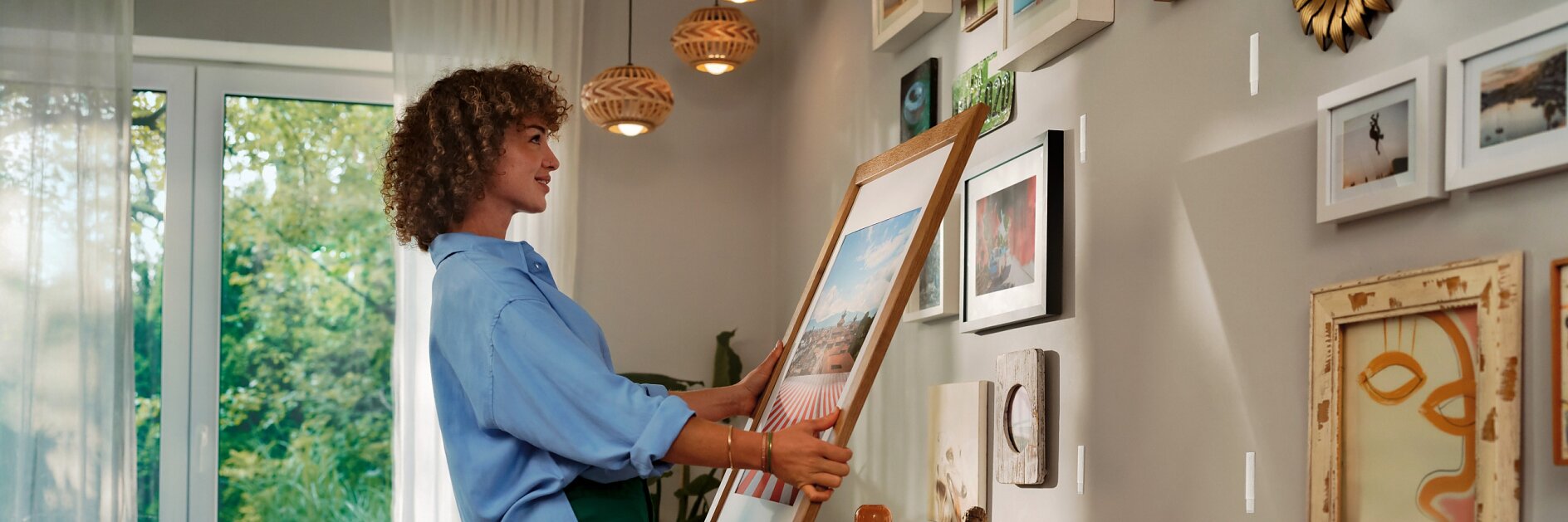 A person in a blue shirt and green pants is hanging a framed picture on a wall using tesa tape. The room is decorated with multiple artworks and has a wooden desk with various items. A window with sheer curtains is open to a view of greenery outside. (This text has been generated by AI)
