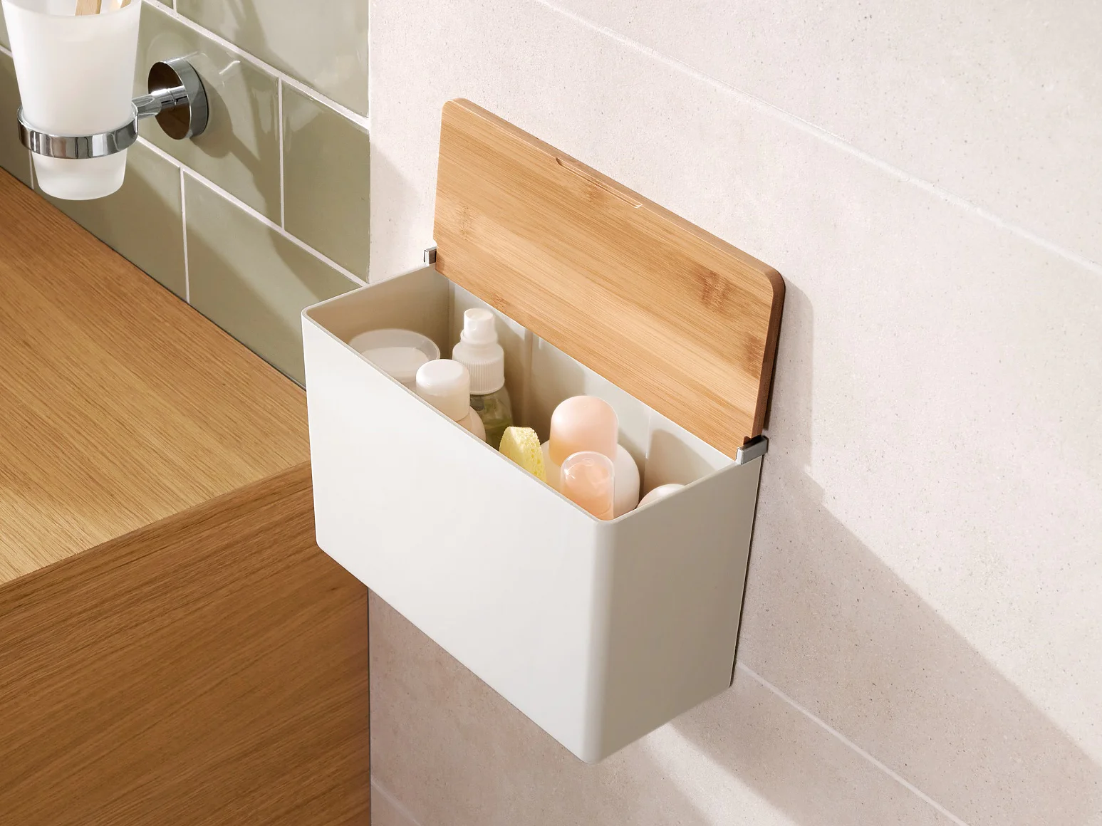 A white rectangular wall-mounted storage box with a wooden lid is open, revealing various bottles inside. It is attached to a bathroom wall using tesa tape, near a wooden countertop with a bowl sink and a tray holding jewelry. A towel hangs to the right. (This text has been generated by AI)