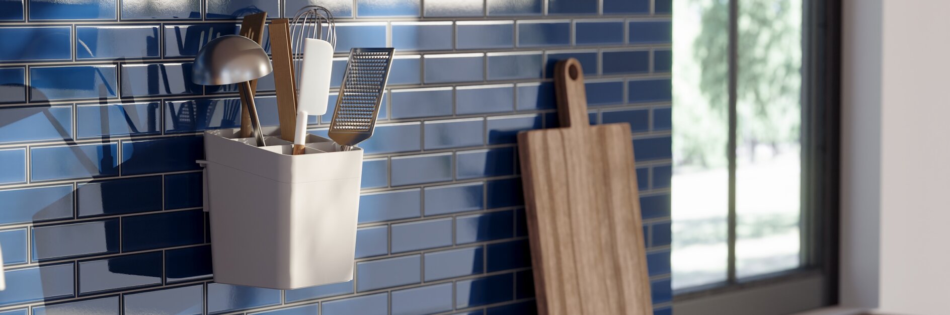 A kitchen wall with glossy blue subway tiles features a hanging white container holding utensils such as a spatula and whisk, all secured with tesa tape. A wooden cutting board leans against the wall. A window to the right lets in natural light. (This text has been generated by AI)