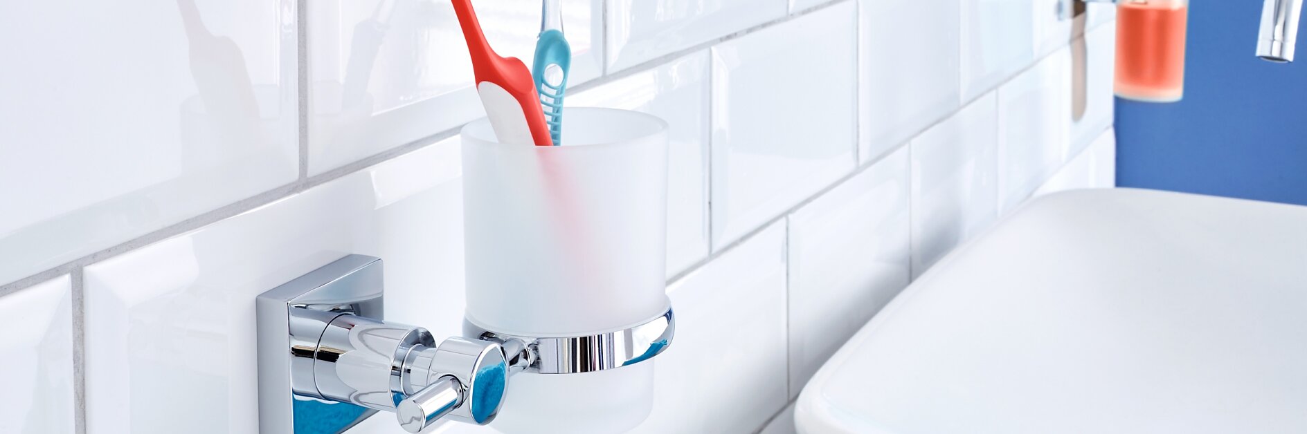 Une salle de bain avec un mur carrelé blanc, un porte-gobelet en verre dépoli fixé au mur contenant une brosse à dents rouge et une bleue, et un lavabo blanc à proximité. Une serviette bleue est posée sur le bord du lavabo, avec un distributeur de savon liquide visible en arrière-plan. (This text has been generated by AI)