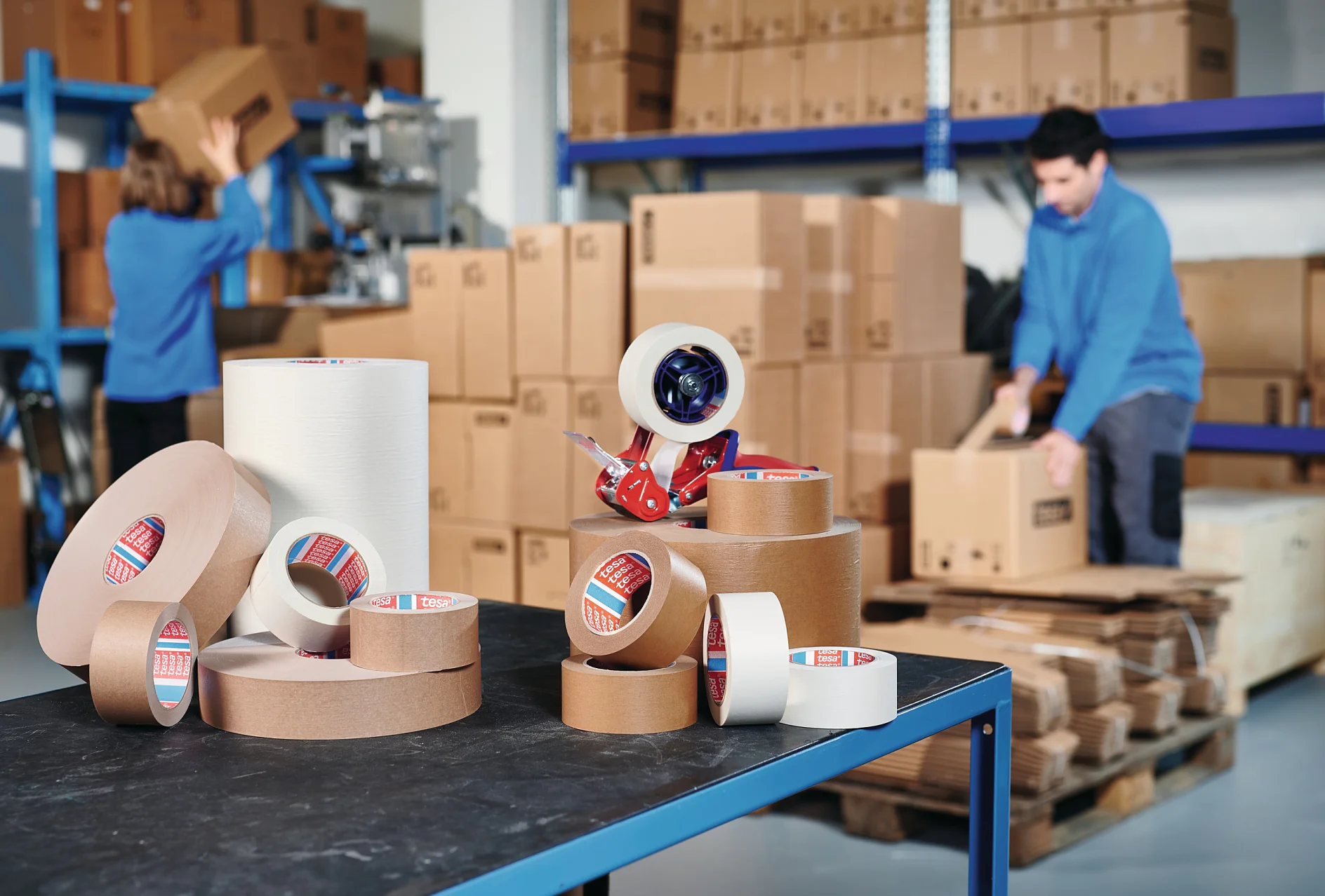 des rubans adhésifs de marque tesa empilés sur une table dans un entrepôt, avec des employés manipulant des cartons à l'arrière-plan (This text has been generated by AI)