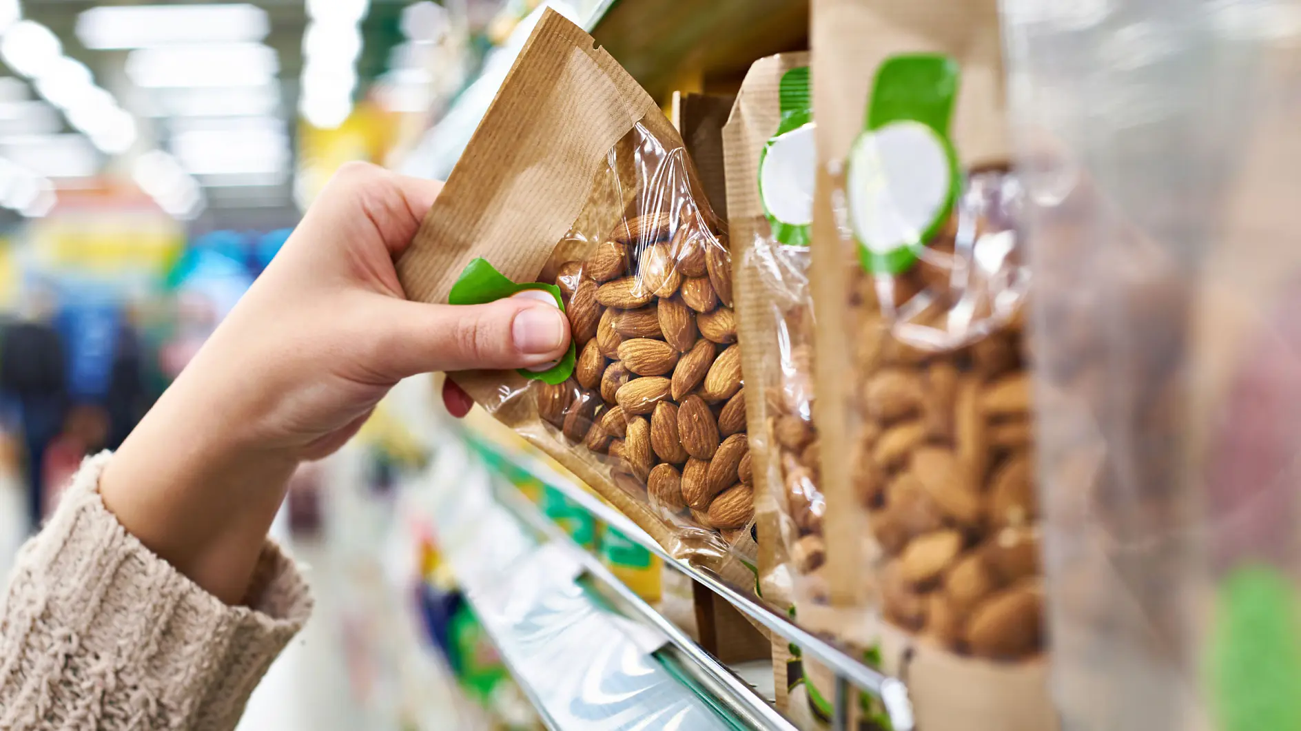 A hand is reaching for a bag of almonds on a store shelf. The almonds are in transparent plastic packaging with a tesa tape backing and a green label. The background shows a brightly lit aisle with various other products displayed.