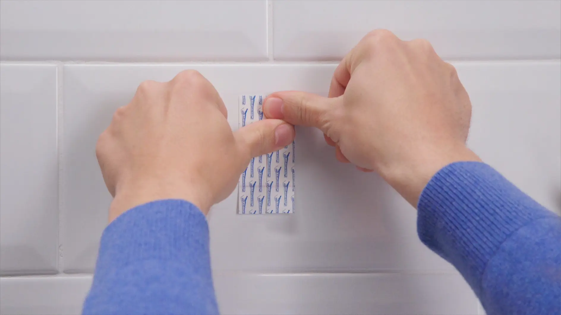 Two hands in blue sleeves are attaching a small tesa tape strip with blue hooks to a white tiled wall. The person is pressing the tesa tape, ensuring it sticks to the surface. (This text has been generated by AI)