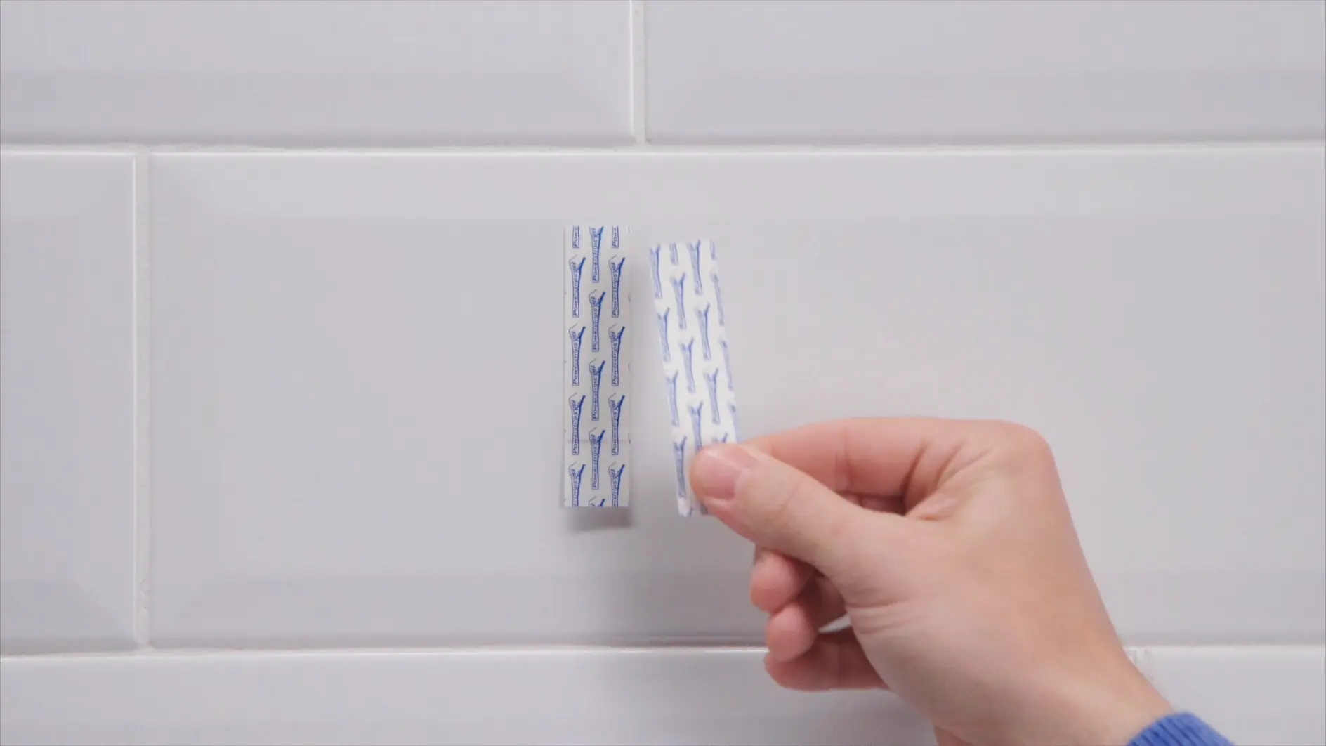 A hand holding a small piece of white tesa tape with blue patterns, positioned against a tiled white wall. Another identical piece is attached to the wall beside it. The background consists of rectangular white tiles. (This text has been generated by AI)