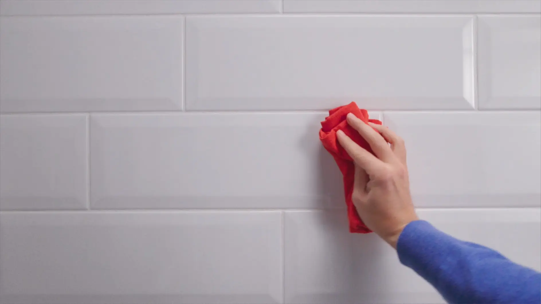 A person wearing a blue sleeve is wiping a white tiled surface with a red cloth. The tiles have a rectangular shape and a glossy finish. The focus is on cleaning activity using tesa tape. (This text has been generated by AI)