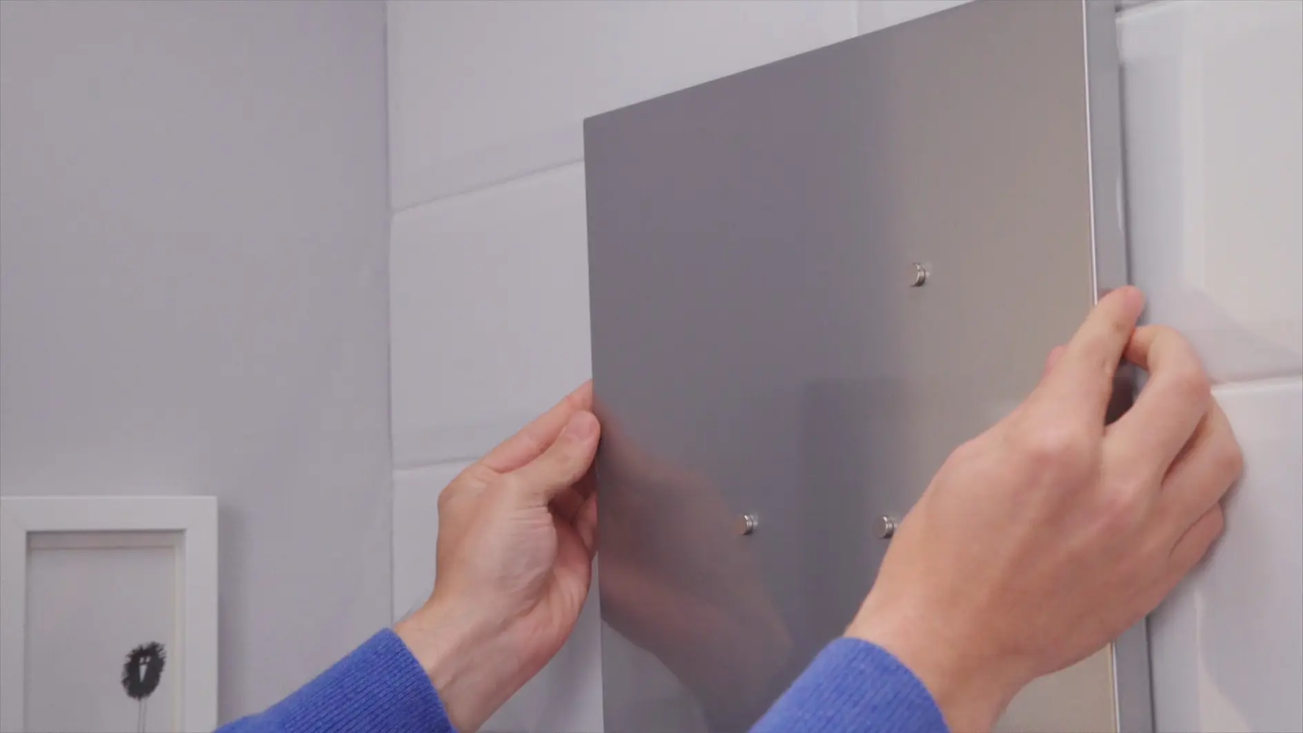 A persons hands are placing a rectangular metal panel onto a white tiled wall, near a small white-framed object. The person is wearing a blue long-sleeved shirt and using tesa tape for the task. (This text has been generated by AI)