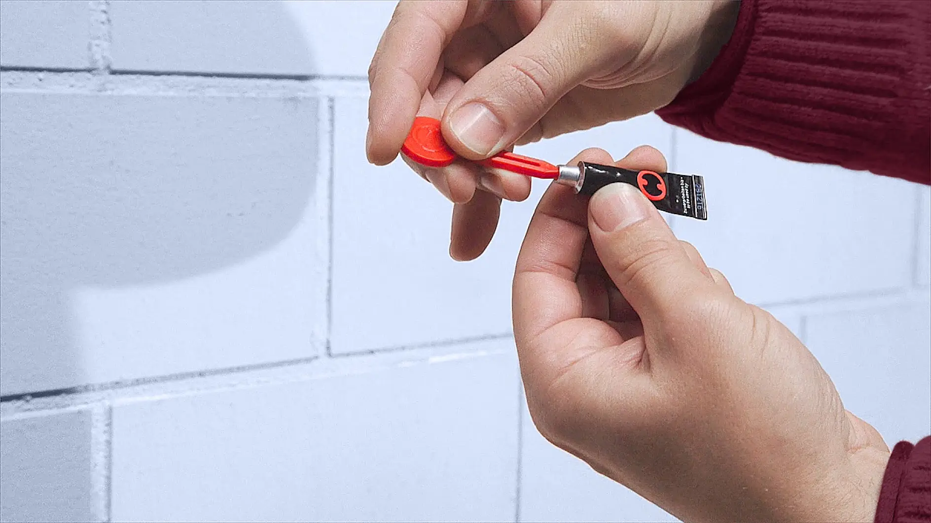 Hands applying glue from a small tube onto a plastic nozzle, in front of a light blue brick wall. The person is wearing a red long-sleeved top. (This text has been generated by AI)