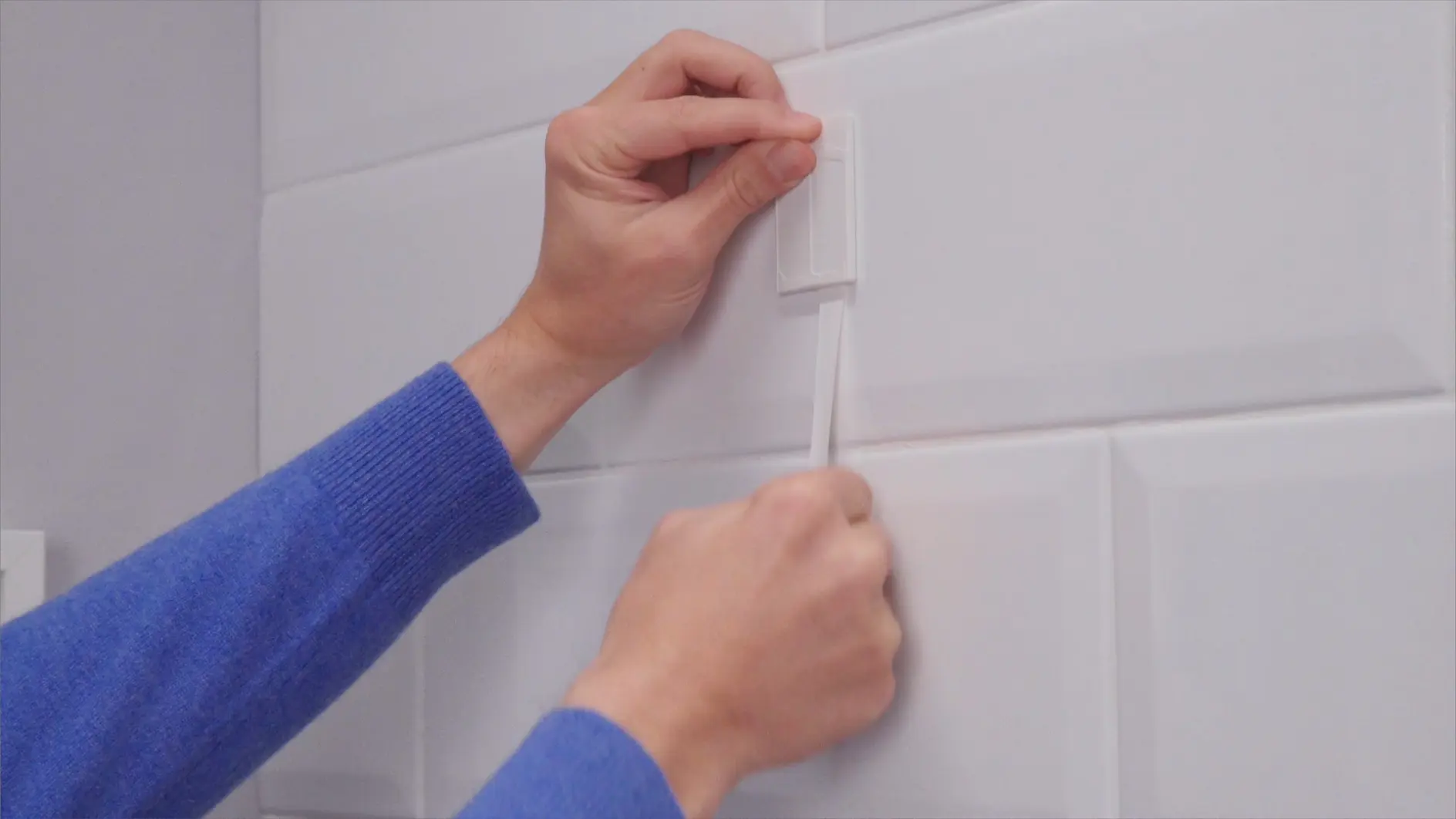 A person in a blue sweater is applying adhesive to a white tiled wall using a rectangular tesa tape strip. They are pressing the tesa tape against the wall with both hands, one holding the strip and the other smoothing it out. (This text has been generated by AI)