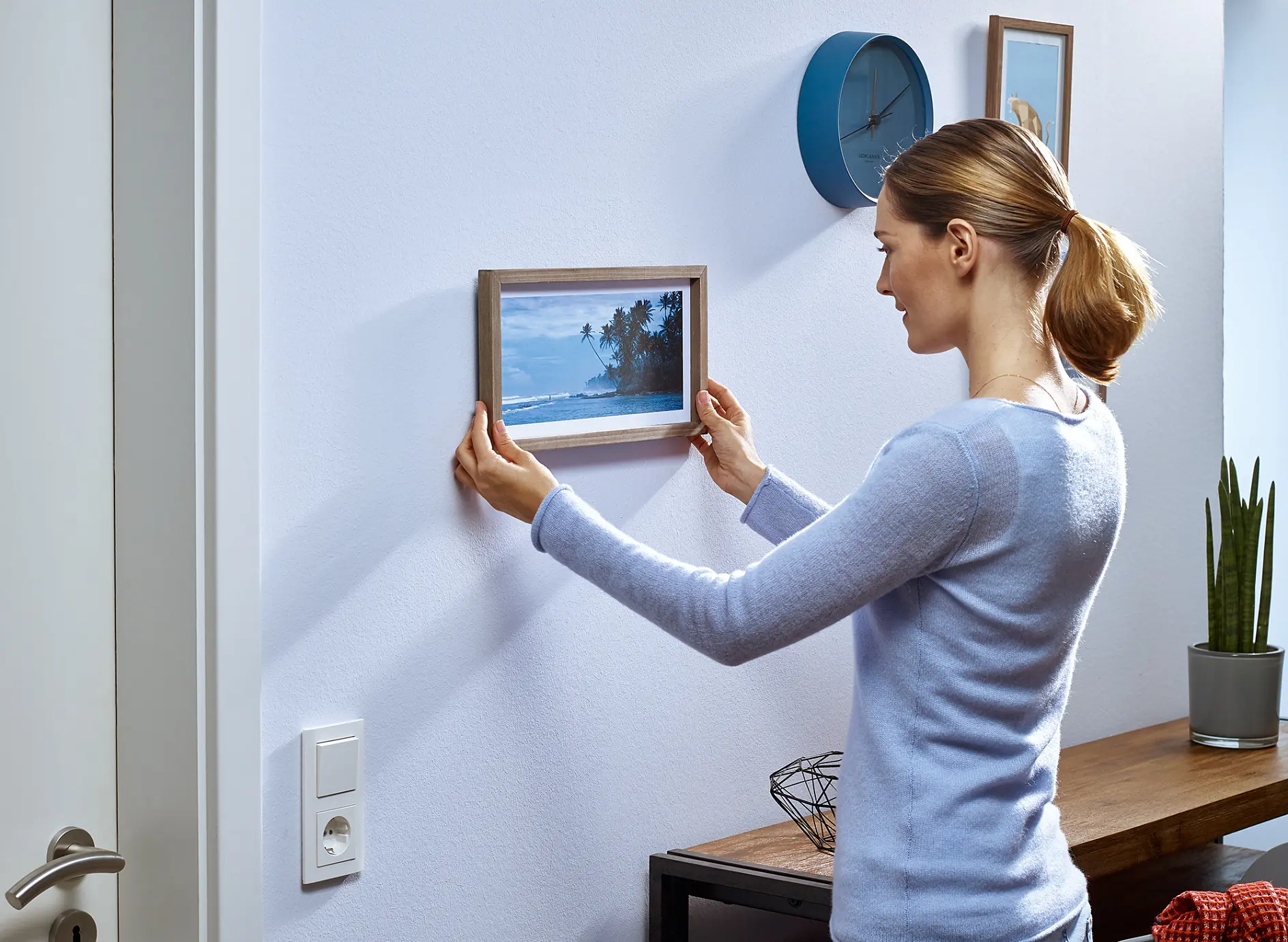 A woman in a light blue sweater is adjusting a framed picture of a beach scene on a white wall with the help of tesa tape. A modern clock and a small plant are on a console table nearby. A light switch and a door are partially visible. (This text has been generated by AI)