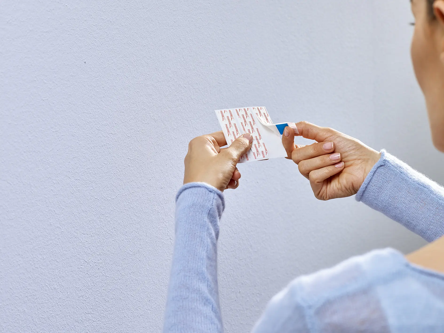 A person wearing a light blue sweater is holding a tesa tape adhesive strip. They are peeling off the backing to prepare it for application against a plain white wall. The focus is on the hands and the strip. (This text has been generated by AI)
