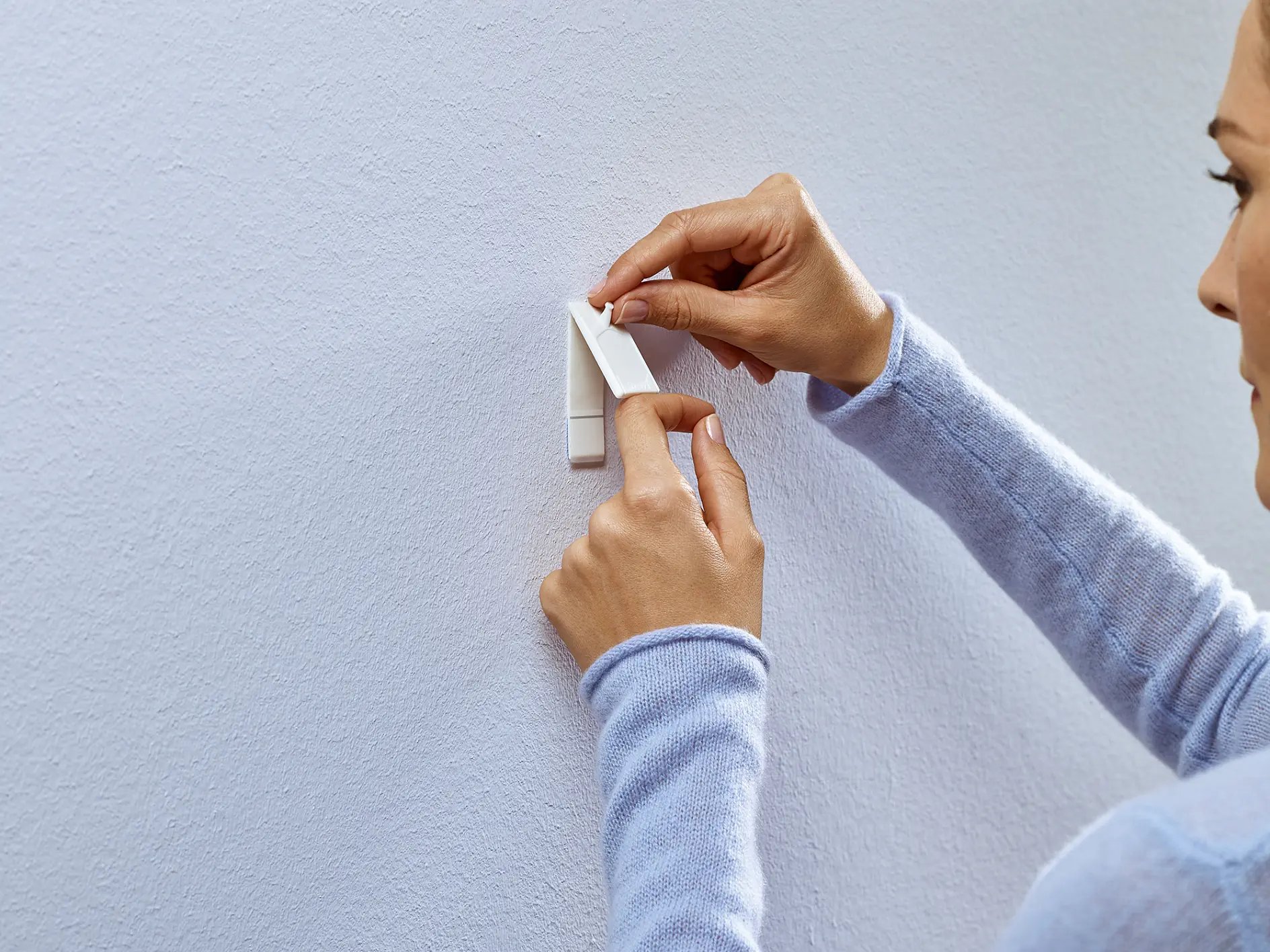 A person wearing a light blue sweater is attaching a white adhesive hook to a smooth, light-colored wall using tesa tape. They are pressing the top of the hook with one hand while holding the base steady with the other. The face is partially visible. (This text has been generated by AI)