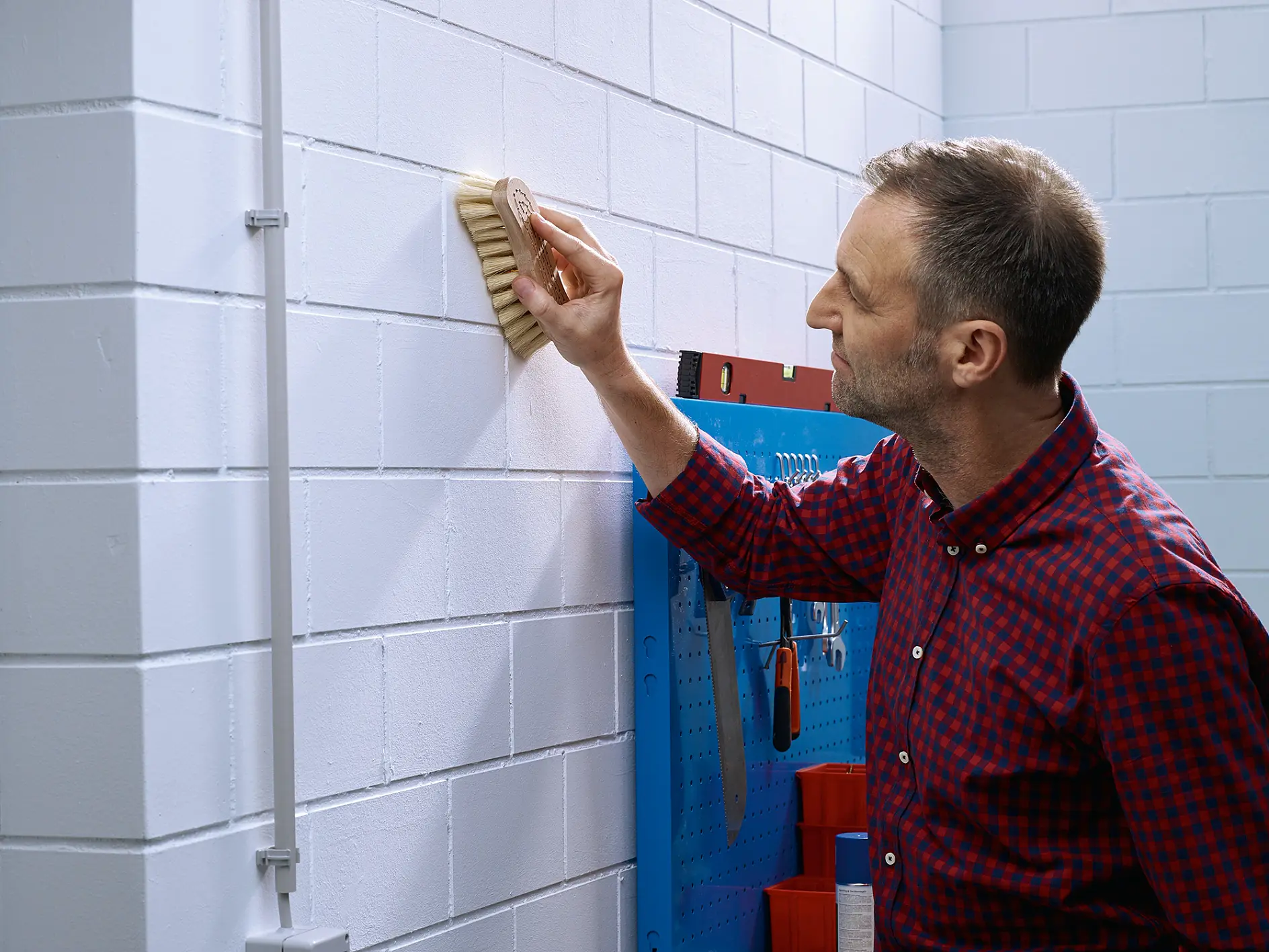 A man in a red plaid shirt is cleaning a white brick wall with a brush in a workshop setting. A blue tool rack with various hand tools and rolls of tesa tape is visible in the background. (This text has been generated by AI)