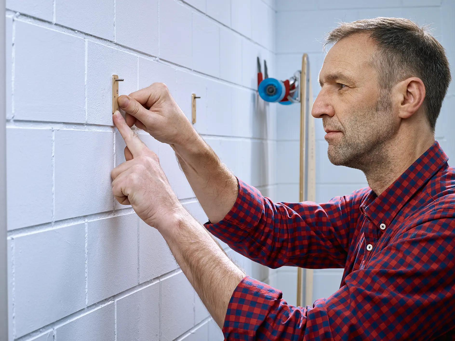 A man in a red plaid shirt is aligning pegs on a white brick wall using tesa tape. Tools are visible in the background on an organizer. The scene appears to be in a workshop or garage, and the man is focused on his task. (This text has been generated by AI)